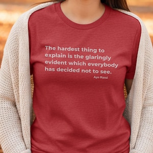 Pode incluir: Camiseta vermelha com texto branco citando Ayn Rand: "A coisa mais difícil de explicar é o que é tão evidente que todos decidiram não ver." A camiseta é usada com um cardigan creme.