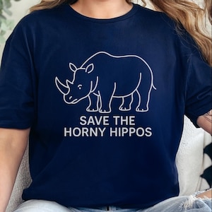 Puede incluir: Camiseta azul marino con un contorno blanco de un rinoceronte y el texto "SAVE THE HORNY HIPPOS". La camiseta tiene cuello redondo y está hecha de tela suave.