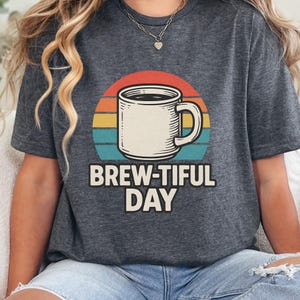 Könnte beinhalten: Dunkelgraues T-Shirt mit einer Kaffeetassen-Grafik und dem Text "BREW-TIFUL DAY". Das Design zeigt eine Kaffeetasse vor einem Retro-Sonnenuntergang mit roten, orangefarbenen, gelben und türkisfarbenen Streifen. Ein lässiges T-Shirt.