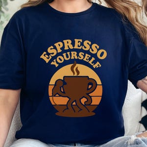 May include: Navy blue t-shirt featuring the text "ESPRESSO YOURSELF" in gold above a brown coffee cup graphic. The coffee cup has legs and arms, set against an orange and yellow sunset design.