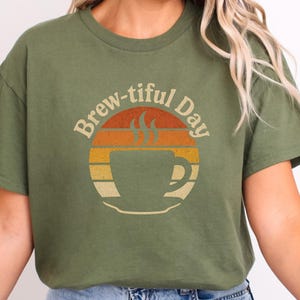 May include: Olive green t-shirt with a retro design. The graphic features a coffee cup silhouette inside a circular design with orange, yellow, and beige stripes. The text "Brew-tiful Day" arches above the cup.