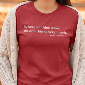 May include: A red t-shirt featuring the quote "Where all think alike, no one thinks very much." by Walter Lippmann in white text. The person is wearing a cream cardigan.