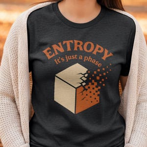 May include: Dark gray t-shirt with the word "ENTROPY" in orange, above the phrase "It's just a phase." A geometric cube design is partially broken apart, with some sections dissolving into orange pixels. The shirt is a casual wear item.