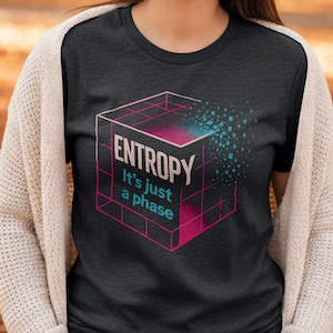 May include: Dark grey t-shirt with a geometric cube design. The text reads "ENTROPY It's just a phase" in white and pink. The design features a pink and blue gradient and a pixelated effect, creating a modern aesthetic.