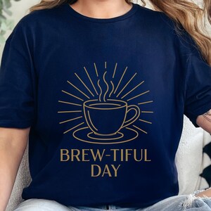 Peut inclure: T-shirt bleu marine avec un motif doré d'une tasse de café fumante entourée de rayons de soleil. Le texte "BREW-TIFUL DAY" est imprimé en dessous. Vêtement décontracté.