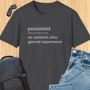 May include: Dark grey t-shirt with white text that reads "pessimist [pes-uh-mist] noun an optimist who gained experience"