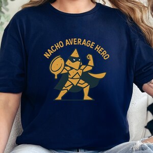 Puede incluir: Camiseta azul marino con un diseño dorado de un superhéroe nacho sosteniendo un plato de nachos. El texto "NACHO AVERAGE HERO" está arqueado sobre la imagen en dorado.