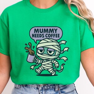 Op de afbeelding: Groen t-shirt met een cartoon mummy ontwerp. De mummie, gewikkeld in verband, houdt een koffiekopje vast met gemorste koffie. Een tekstballon boven de mummie leest "MUMMY NEEDS COFFEE". Het ontwerp gebruikt zwart, wit, bruin en grijs.