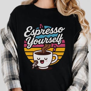 Puede incluir: Camiseta negra con la frase "Espresso Yourself" en escritura blanca sobre una taza de café de dibujos animados con un micrófono. El diseño incluye un fondo colorido estilo puesta de sol con notas musicales.