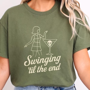 May include: Olive green t-shirt featuring a white graphic of a woman, a martini glass with a skull, and the text "Swinging 'til the end". The design is centered on the front of the shirt.