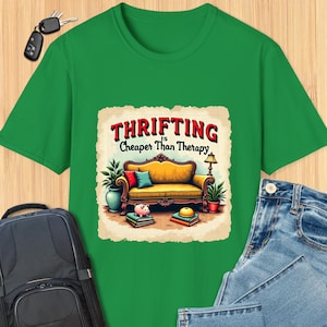 May include: Green t-shirt with a vintage-style illustration of a yellow couch with pillows, a piggy bank, and books. The text "Thrifting is Cheaper Than Therapy" is printed on the shirt.