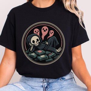 May include: Black t-shirt with a cartoon Grim Reaper resting on video game cartridges, with three pink ghosts. The Grim Reaper is in a black hooded robe and has a skull face. The design is in a circular frame.