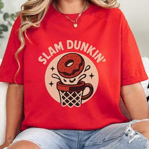 Pode incluir: Camiseta vermelha com um design gráfico que apresenta um donut mergulhando em uma xícara de café em forma de cesta de basquete. O texto "SLAM DUNKIN'" está arqueado acima do donut. O design é em tons de marrom e creme.