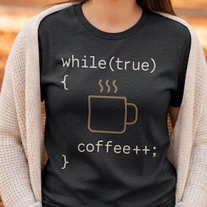 Op de afbeelding: Donkergrijs t-shirt met de tekst "while(true) { coffee++; }" in lichtbeige. Er is ook een afbeelding van een koffiemok. Dit shirt is een casual kledingstuk.