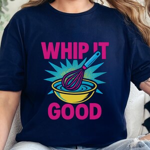 Puede incluir: Camiseta azul marino con la frase "WHIP IT GOOD" en rosa. Un batidor se muestra sobre un cuenco amarillo con líquido azul, rodeado de un diseño de explosión turquesa. Es una prenda de vestir informal.