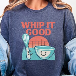 Puede incluir: Camiseta azul marino con la frase "WHIP IT GOOD" en letras naranjas. Debajo del texto hay una ilustración de dibujos animados de un batidor, un cuenco y una puesta de sol. El batidor y el cuenco tienen caras sonrientes. Una camiseta divertida para panaderos.