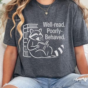 Puede incluir: Camiseta gris oscuro con un gráfico blanco de un mapache leyendo un libro y una pila de libros. El texto "Well-read. Poorly-Behaved." está impreso en la camiseta. La camiseta es de estilo casual.