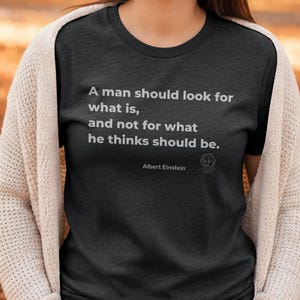 May include: A charcoal grey t-shirt featuring the quote "A man should look for what is, and not for what he thinks should be." by Albert Einstein. The text and Einstein's illustration are in white.