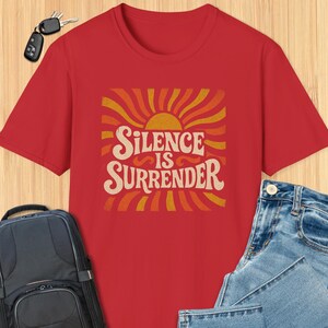 May include: A red t-shirt featuring the phrase "Silence is Surrender" in a retro style, with an orange and yellow sunburst design. The shirt is shown with blue jeans and a black backpack.