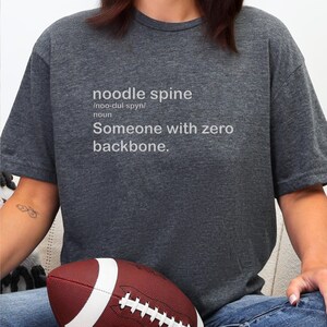May include: Dark gray t-shirt with the text "noodle spine" and the definition "Someone with zero backbone." A brown and white football is in the foreground.
