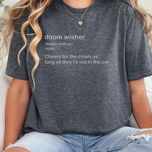 May include: Dark gray t-shirt with the text "doom wisher" and its definition. The definition reads "Cheers for the crash as long as they're not in the car." The shirt is worn by a person with blonde hair and a silver necklace.