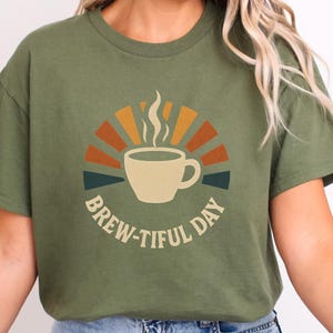 Peut inclure: T-shirt vert olive avec un motif de tasse à café et de vapeur, sur un fond de soleil orange, jaune et sarcelle. Le texte "BREW-TIFUL DAY" est en arc de cercle sous la tasse. Un t-shirt décontracté et confortable.