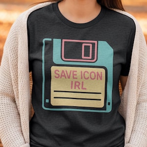 May include: Dark gray t-shirt featuring a retro-style floppy disk graphic in teal, pink, and yellow. The text "SAVE ICON IRL" is printed on the disk. The shirt is a crew neck.