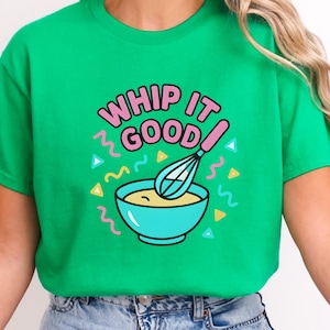 Puede incluir: Camiseta verde con la frase "Whip It Good" en letras rosas. El diseño incluye un cuenco azul con un batidor y líneas onduladas y triángulos de colores. La camiseta es de cuello redondo.