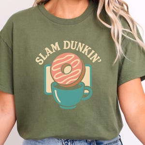 May include: An olive green t-shirt featuring a graphic of a pink iced donut above a teal coffee cup. The text "SLAM DUNKIN'" is displayed above the donut in a retro font.