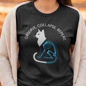 Puede incluir: Camiseta gris oscuro con una silueta de gato blanco y un diseño esquelético azul. El texto "OBSERVE. COLLAPSE. REPEAT." está arqueado sobre el gato. La camiseta es de cuello redondo.
