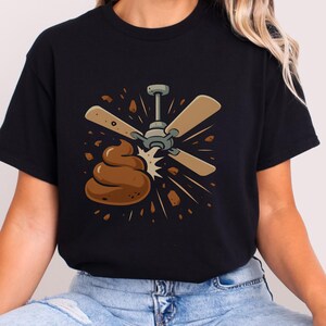 May include: Black t-shirt featuring a humorous graphic of a ceiling fan with brown blades and a pile of poop. The design includes brown debris and a starburst effect, creating a comical and eye-catching image.