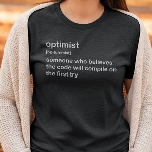 Könnte beinhalten: Dunkelgraues T-Shirt mit der Definition von "Optimist" in Weiß aufgedruckt. Die Definition lautet: "jemand, der glaubt, dass der Code beim ersten Versuch kompiliert wird." Das T-Shirt hat einen Rundhalsausschnitt.