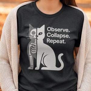 May include: Dark gray t-shirt featuring a white cat design, half skeleton and half solid. The text "Observe. Collapse. Repeat." is printed in white. The shirt is a crew neck style.