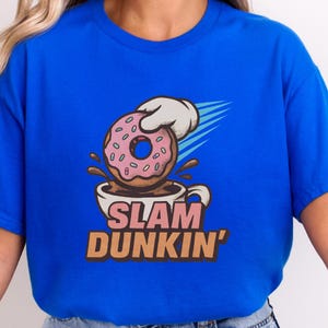 May include: A blue t-shirt featuring a graphic of a pink iced doughnut being dunked into a coffee cup. The text "SLAM DUNKIN'" is below the cup in brown and pink letters. The doughnut is held by a white gloved hand.