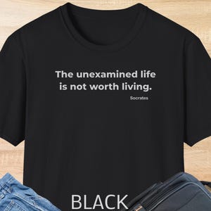 May include: Black t-shirt featuring the quote "The unexamined life is not worth living" by Socrates in white text. The word "BLACK" is printed at the bottom of the shirt.