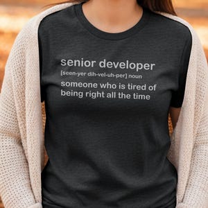 May include: A dark grey t-shirt featuring the words "senior developer" and its pronunciation, followed by a definition: "someone who is tired of being right all the time." The shirt is worn with a cream cardigan.