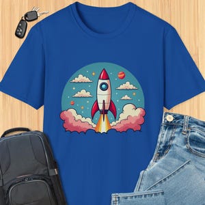 May include: A blue t-shirt with a cartoon rocket ship launching into space. The rocket is red and white with a blue flame. The background is a circle with clouds, stars, and planets.