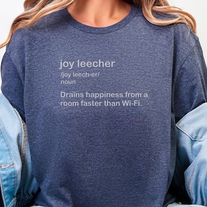 May include: A navy blue t-shirt featuring the definition of "joy leecher" in white text. The definition describes someone who drains happiness from a room faster than Wi-Fi. The shirt's design is simple and direct.