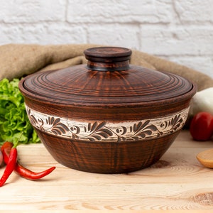 May include: A brown ceramic pot with a lid, featuring a white floral design around the rim. The pot is sitting on a wooden surface with red peppers and green lettuce in the background.