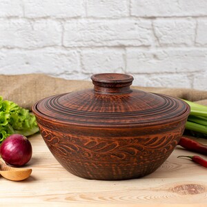 May include: A brown ceramic pot with a lid, featuring a decorative floral design. The pot is sitting on a wooden surface with vegetables and a wooden spoon.