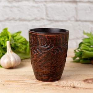 Może przedstawiać: Brązowy kubek ceramiczny z rzeźbionym wzorem kwiatowym. Kubek stoi na drewnianej powierzchni.