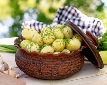 May include: A brown ceramic pot with a lid filled with boiled potatoes and fresh dill. The pot is decorated with a floral pattern. The potatoes are in a rustic setting on a wooden table.