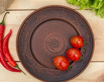 Pode incluir: Um prato de cerâmica marrom rústico com um design floral decorativo, com três tomates vermelhos na borda. O prato está sobre uma superfície de madeira com um saco de juta, alface verde e um bulbo de alho.