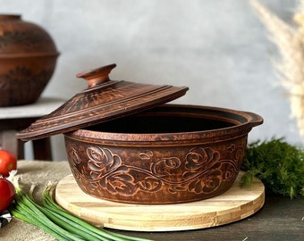 Könnte beinhalten: Eine braune Keramik-Auflaufform mit Deckel, verziert mit einem Blumendesign. Die Form steht auf einem Holzbrett.