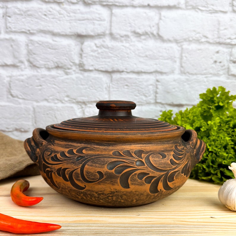 May include: A brown ceramic casserole dish with a lid. The dish features a dark floral pattern and two handles. The background includes a white brick wall, a wooden surface, and some vegetables.