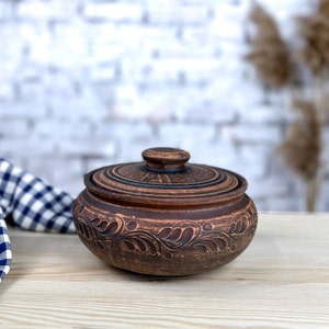 Puede incluir: Una olla de cerámica marrón con tapa. La olla presenta un diseño floral tallado alrededor del exterior. La tapa tiene un pequeño pomo y detalles de círculos concéntricos. La olla se utiliza probablemente para cocinar o almacenar.