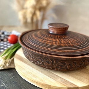 Puede incluir: Una olla de cerámica marrón con tapa, con un diseño floral. La olla está sobre una tabla de cortar de madera.
