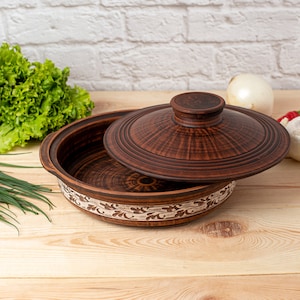 Puede incluir: Una fuente de cerámica marrón con tapa, sobre una superficie de madera clara. El plato tiene una banda decorativa con un diseño floral. Verduras frescas, incluyendo lechuga y una cebolla, están en el fondo. La tapa está parcialmente abierta.