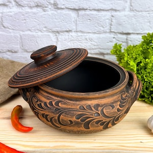 Puede incluir: Una olla de cerámica hecha a mano, abierta, con tapa, con exterior marrón oscuro e interior negro. La olla tiene tallas florales decorativas y dos asas. La tapa está parcialmente abierta. La olla está sobre una superficie de madera.