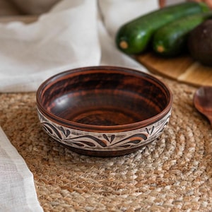 May include: A brown ceramic bowl with a white and black floral design sits on a woven jute placemat. The bowl is empty and ready to be filled.